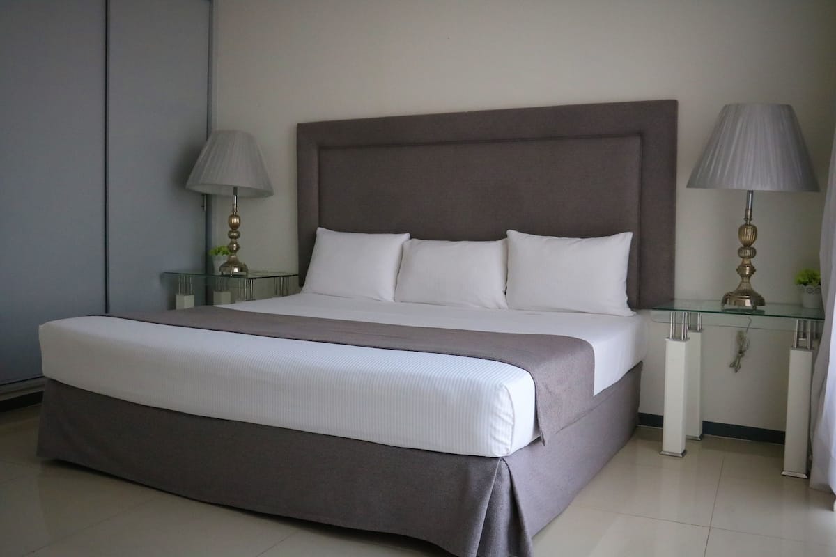 A neatly made bed with a grey headboard and white bedding, two bedside tables with lamps, and a glass side table.