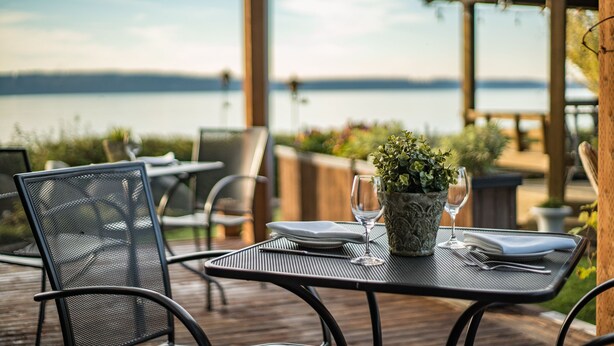 A dining table set outdoors with chairs, a potted plant, and a view of a lake. - Camano Island Inn (Camano Island)
