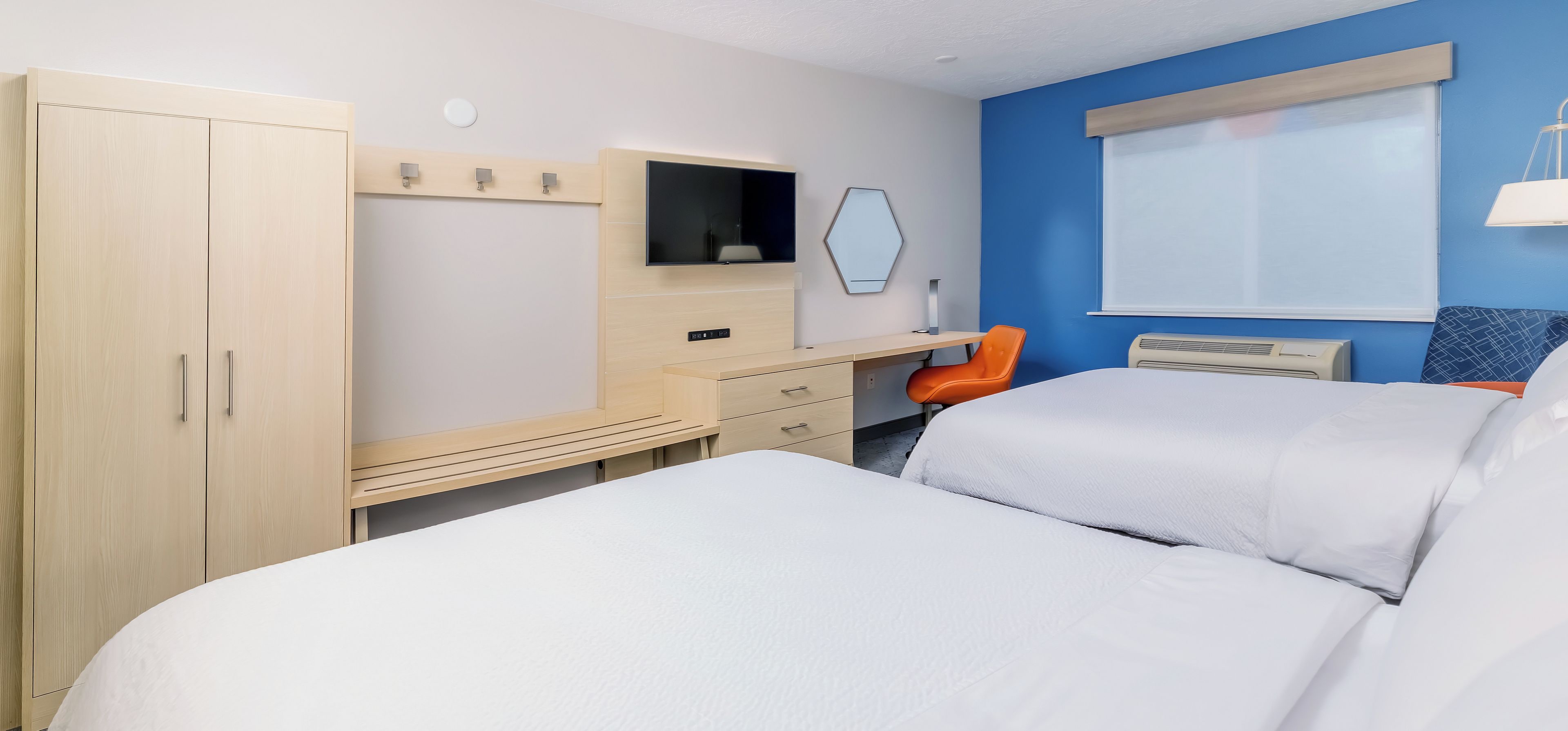 A hotel room with a bed, a desk with a chair, a wardrobe, a TV, and a window with blinds.