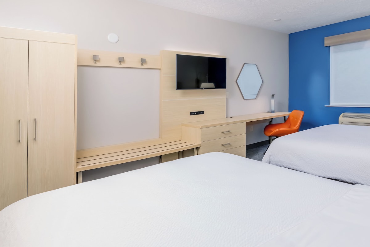 A hotel room with a bed, a desk with a chair, a wardrobe, a TV, and a window with blinds.