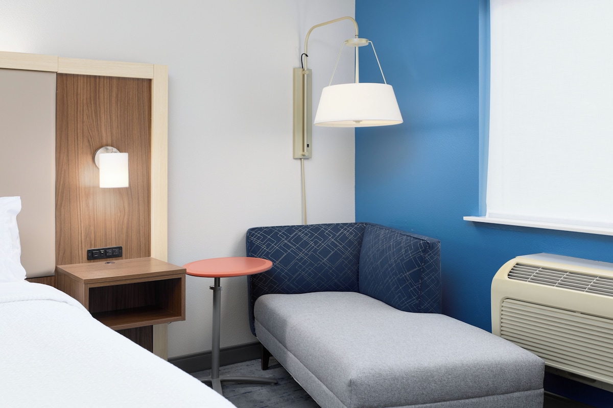 A hotel room with a bed, a bedside table, a small stool, a lamp, and an air conditioning unit.
