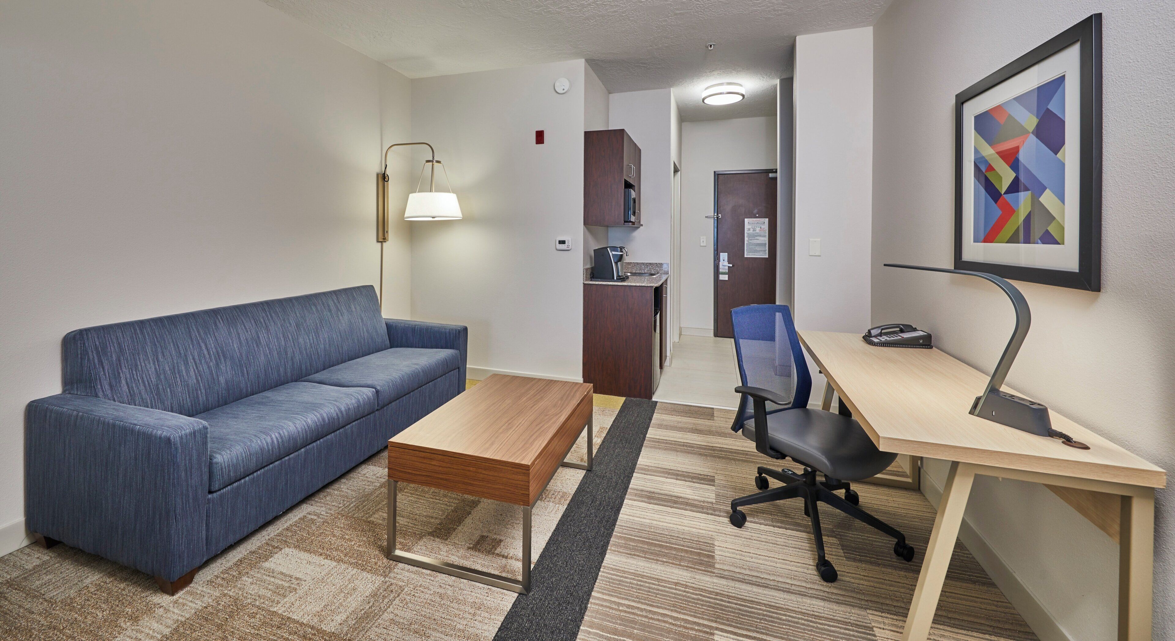 A hotel room with a blue sofa, a wooden coffee table, a desk with a chair, a lamp, and a framed abstract painting.