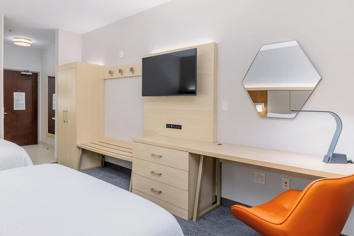 A hotel room with a bed, a desk with a TV, a mirror, and an orange chair.