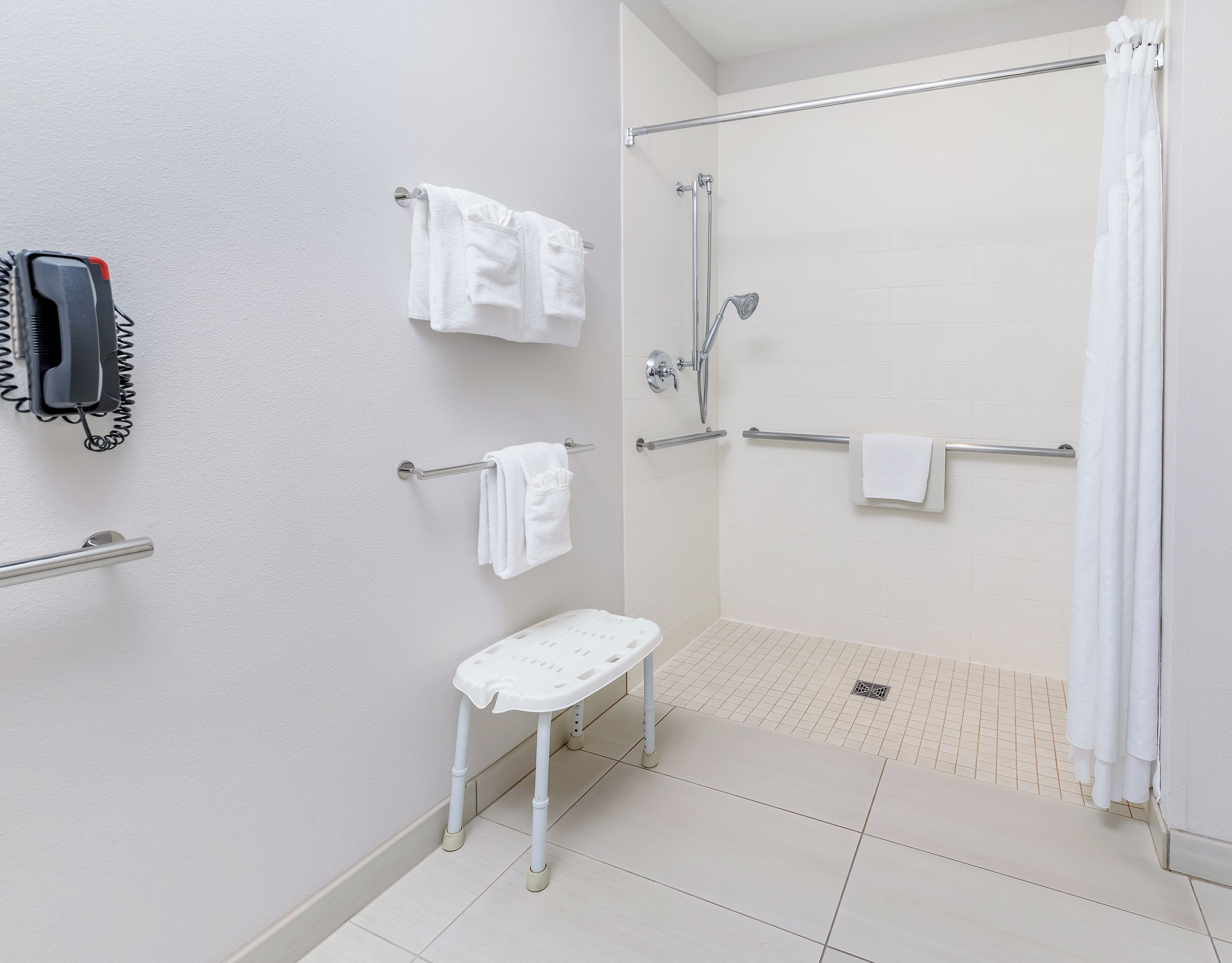 A modern bathroom with a walk-in shower, a toilet with grab bars, a bench, and a phone mounted on the wall.