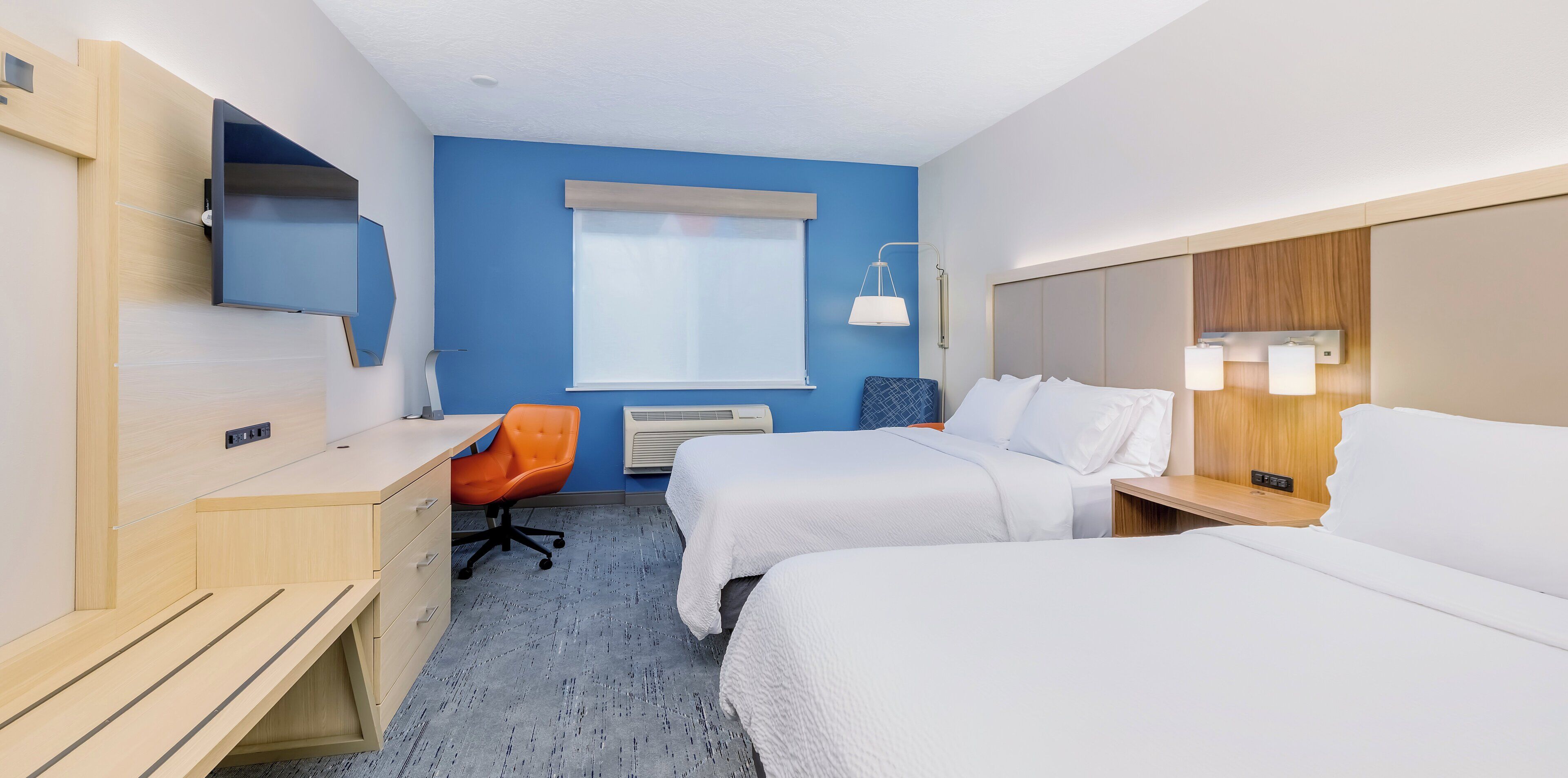 Ein Hotelzimmer mit zwei Betten, einem Schreibtisch, einem orangefarbenen Sessel, einem Fenster mit Jalousien und einem an der Wand befestigten Fernseher.