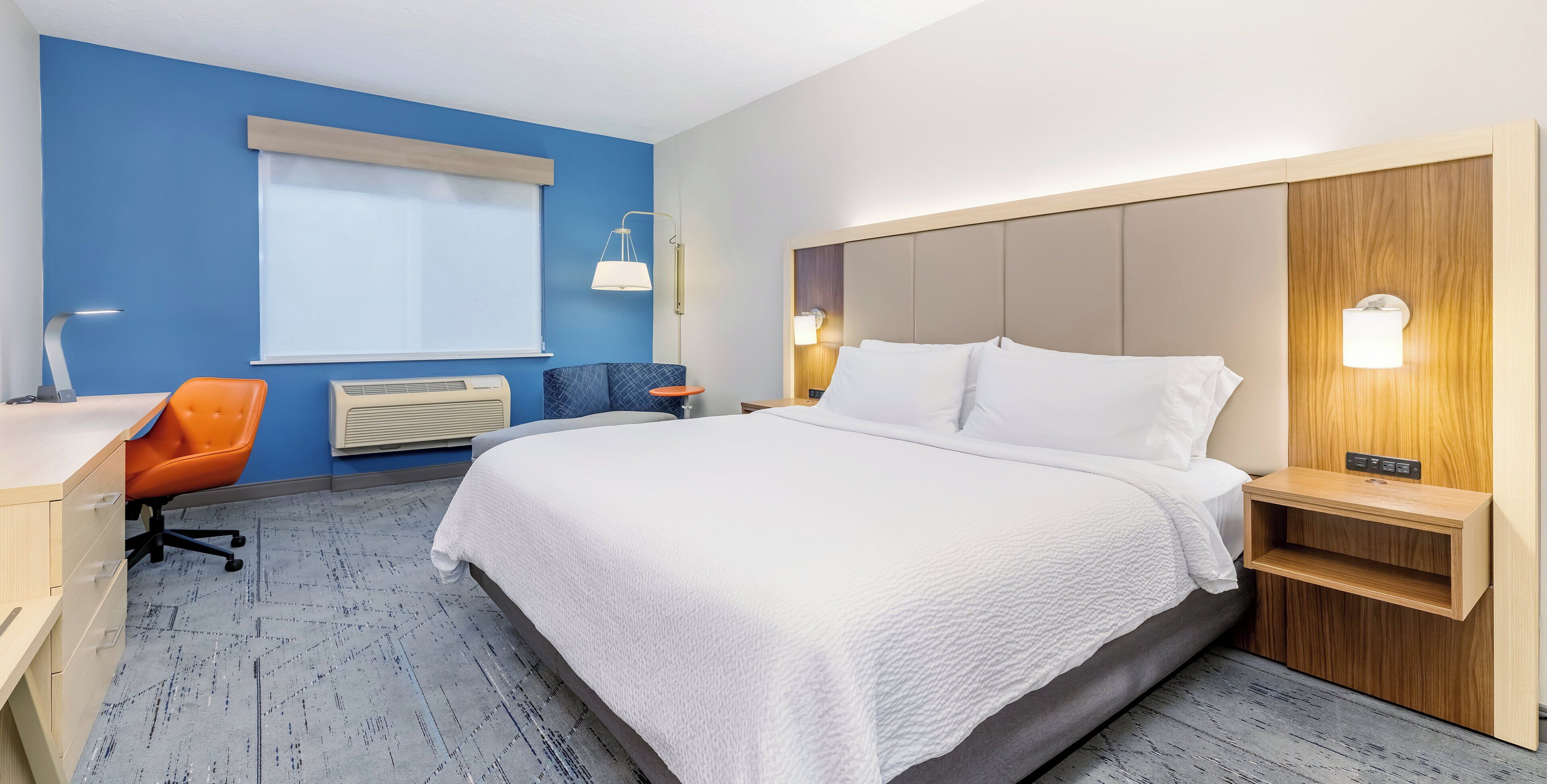Een hotelkamer met een bed, een bureau, een oranje stoel, een blauwe stoel, een raam met zonwering en een wandlamp.