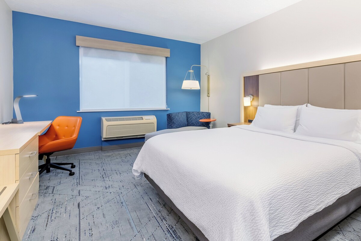 A hotel room with a bed, a desk, an orange chair, a blue chair, a window with blinds, and a wall-mounted lamp.
