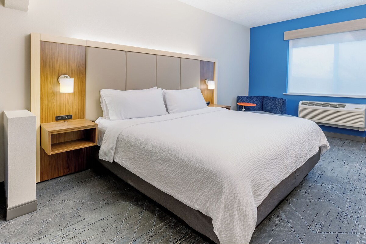 A modern hotel room with a large bed, bedside tables, a nightstand, a window with blinds, and a wall-mounted air conditioner.