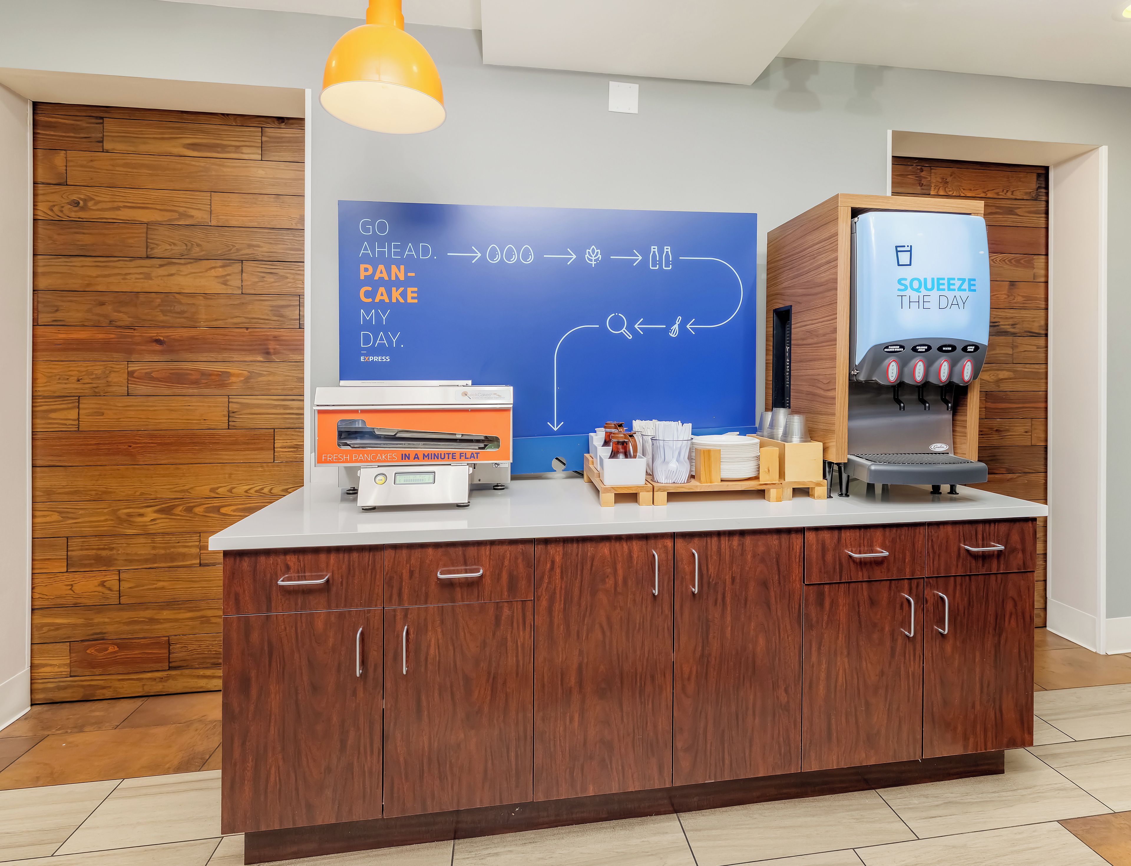 A kitchen area with a flat-screen TV displaying a breakfast menu, a coffee machine, and a wooden cabinet with drawers.