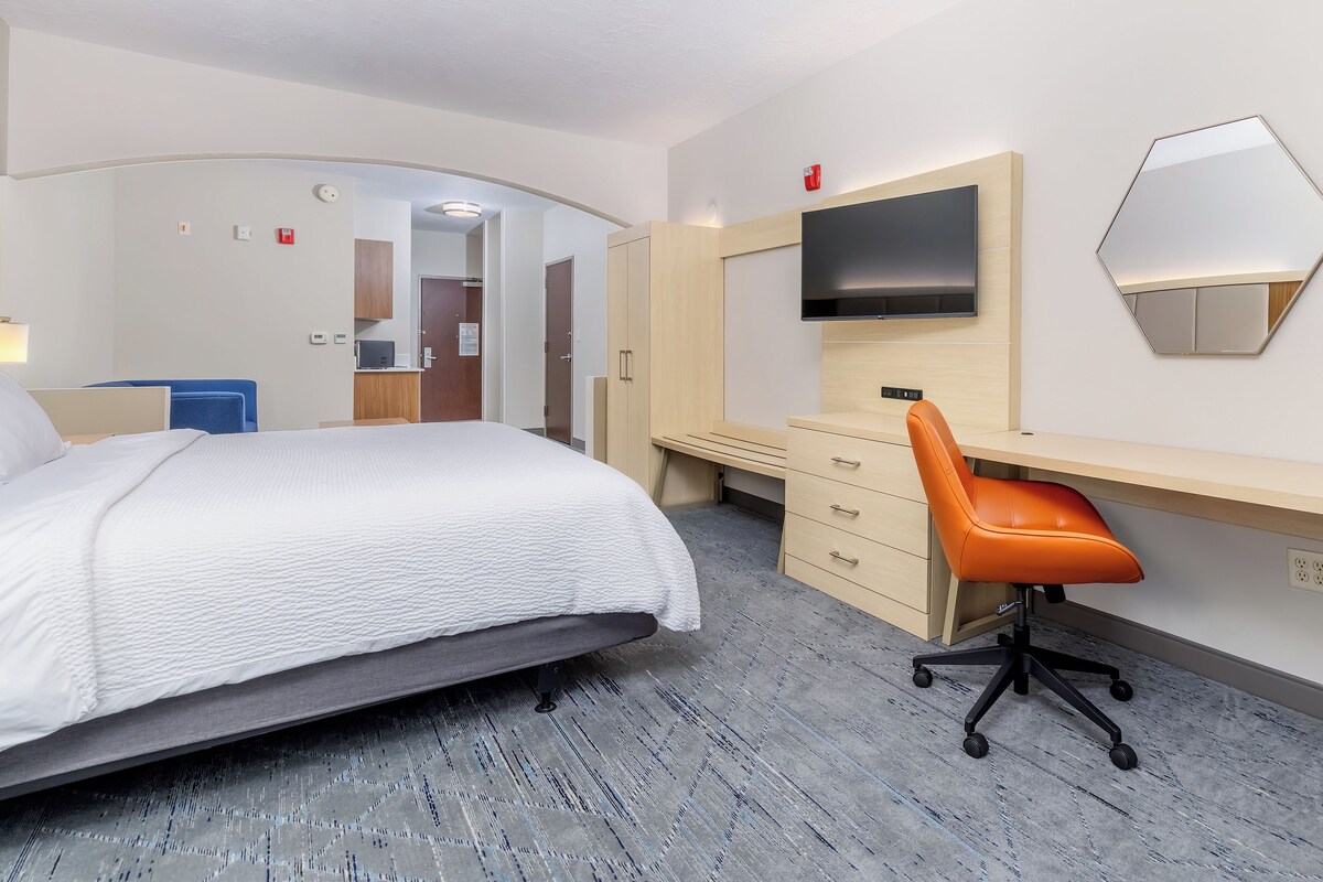 A hotel room with a bed, a desk, an orange chair, a TV, and a mirror.