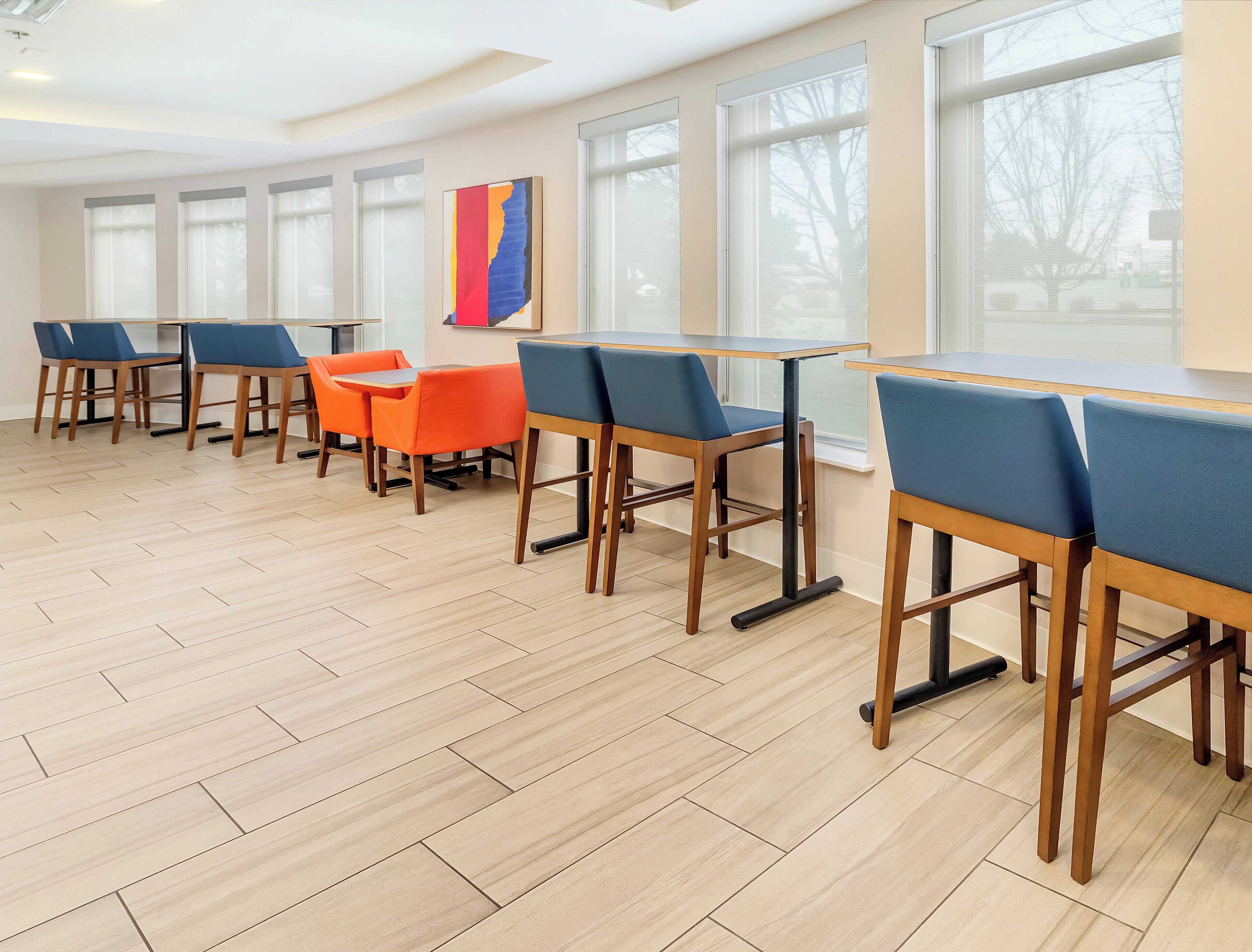 A modern dining area with wooden tables and chairs, large windows, and a herringbone floor pattern.