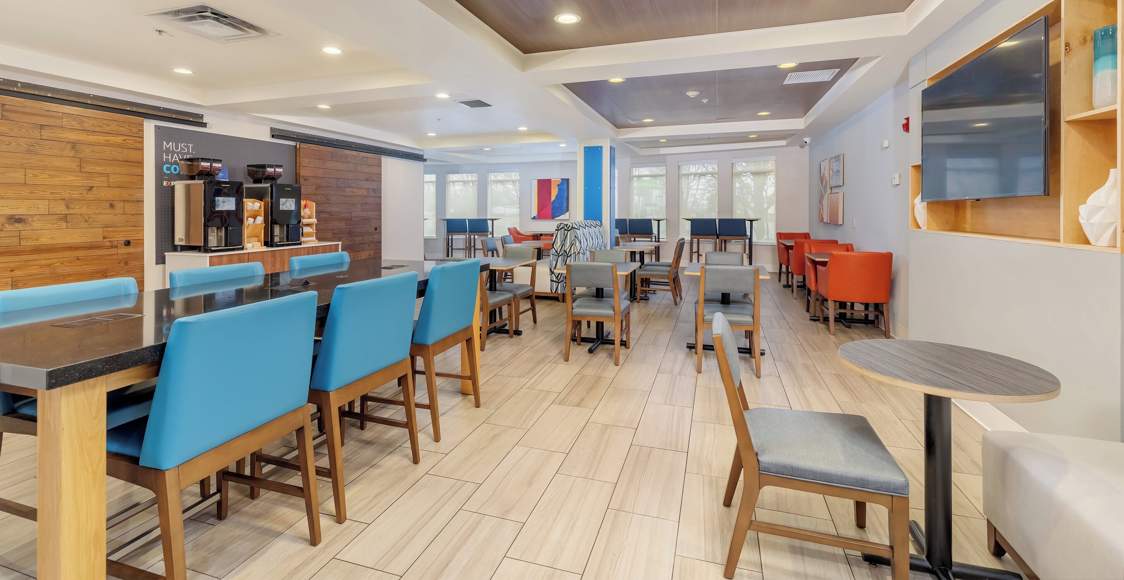 A modern restaurant with wooden flooring, blue and grey chairs, and a flat-screen TV mounted on the wall.