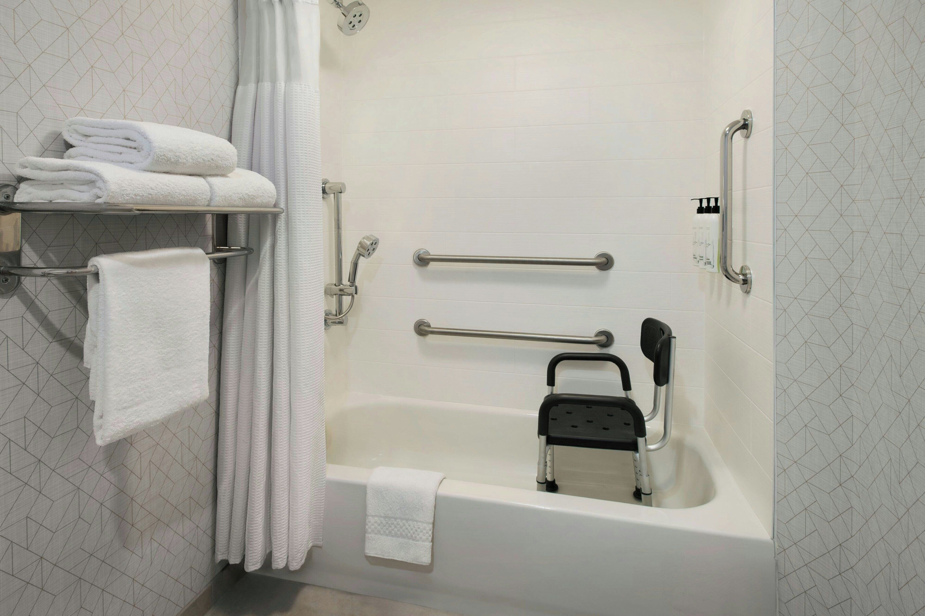 Une salle de bain équipée d’une baignoire, de barres d’appui, d’un siège de douche et de serviettes soigneusement pliées, rangées sur une étagère.
