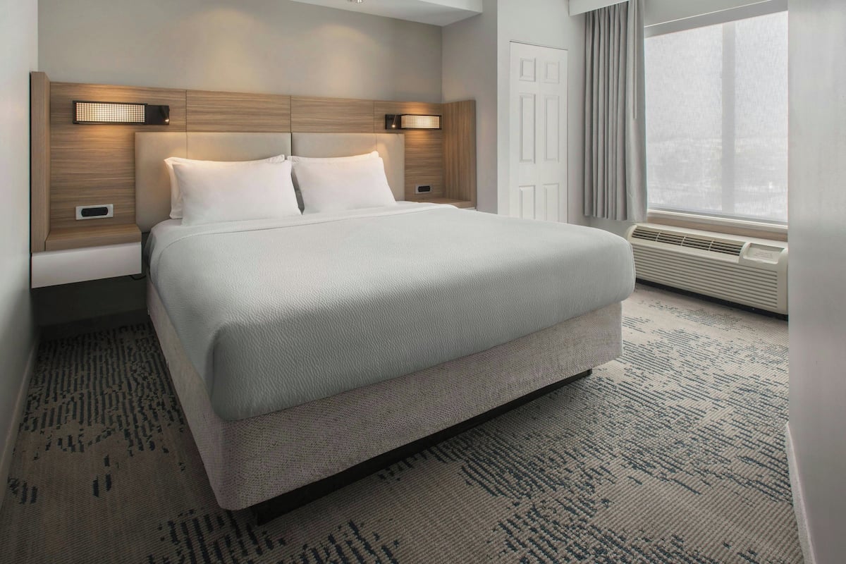 Habitación de hotel con una cama grande, dos mesitas de noche, una lámpara de pared, una ventana con cortinas y un aire acondicionado integrado.