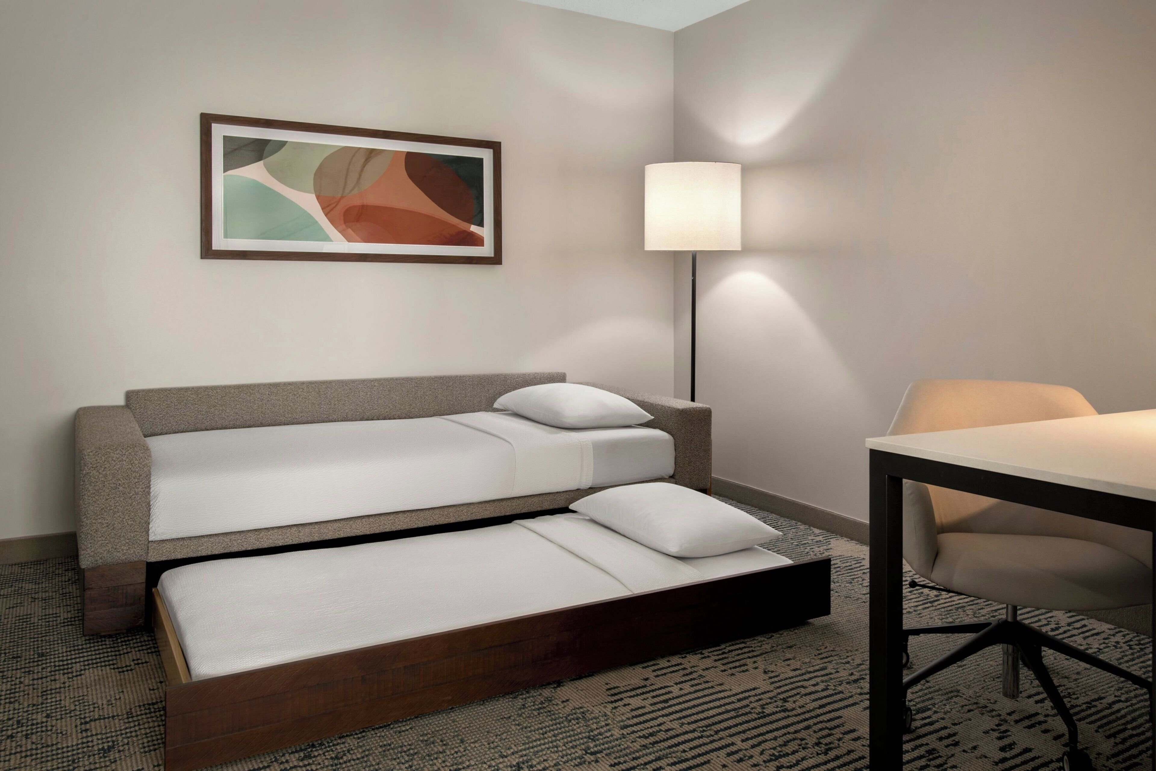 A hotel room with a sofa bed, a desk, a lamp, and a framed abstract painting on the wall.