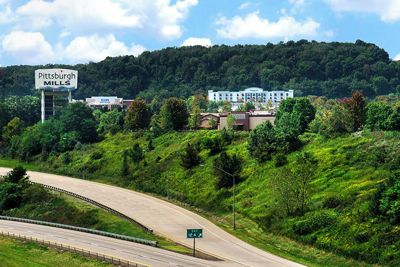 A highway with a sign for Pittsburgh Mills, a large building in the background, and a green road sign indicating Exit 12 A.