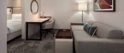 Una habitación de hotel con una cama, un escritorio, una silla, un sofá, una mesita pequeña, una lámpara y un cuadro en la pared.