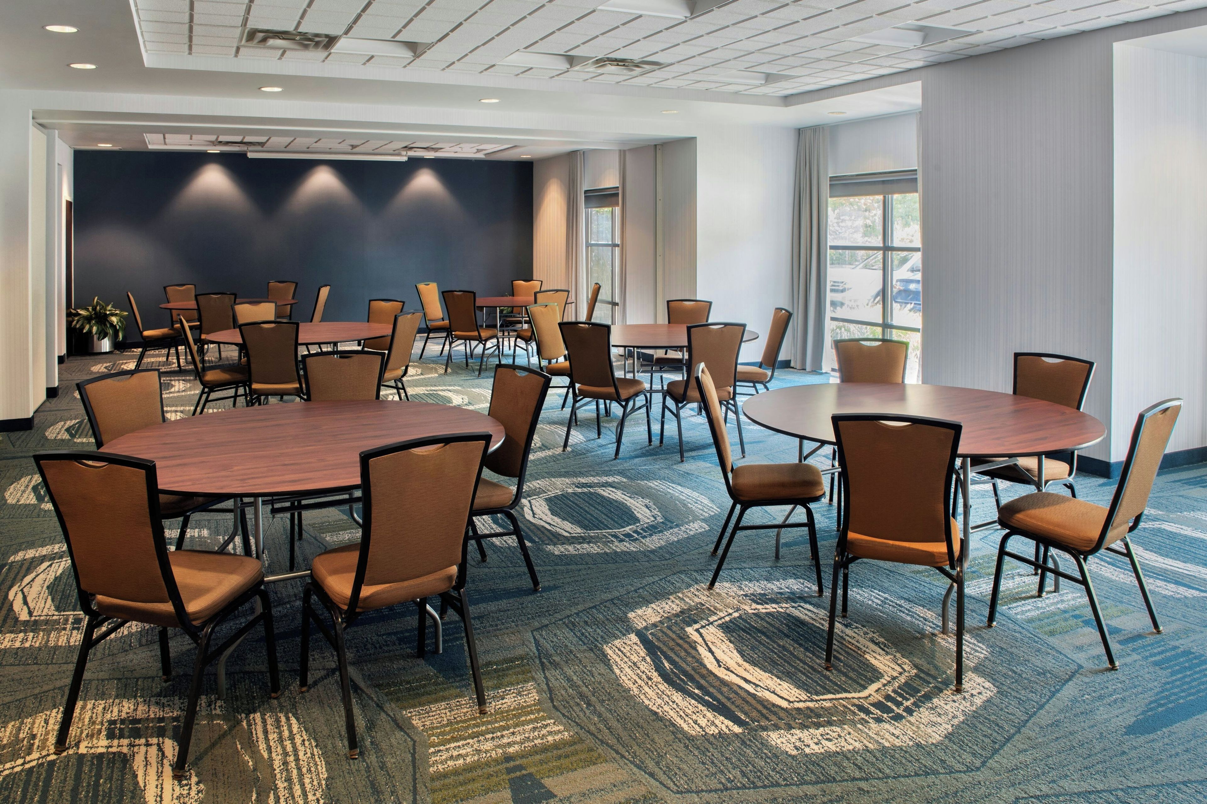 A conference room with multiple tables and chairs, carpeted flooring, and a large window providing natural light.