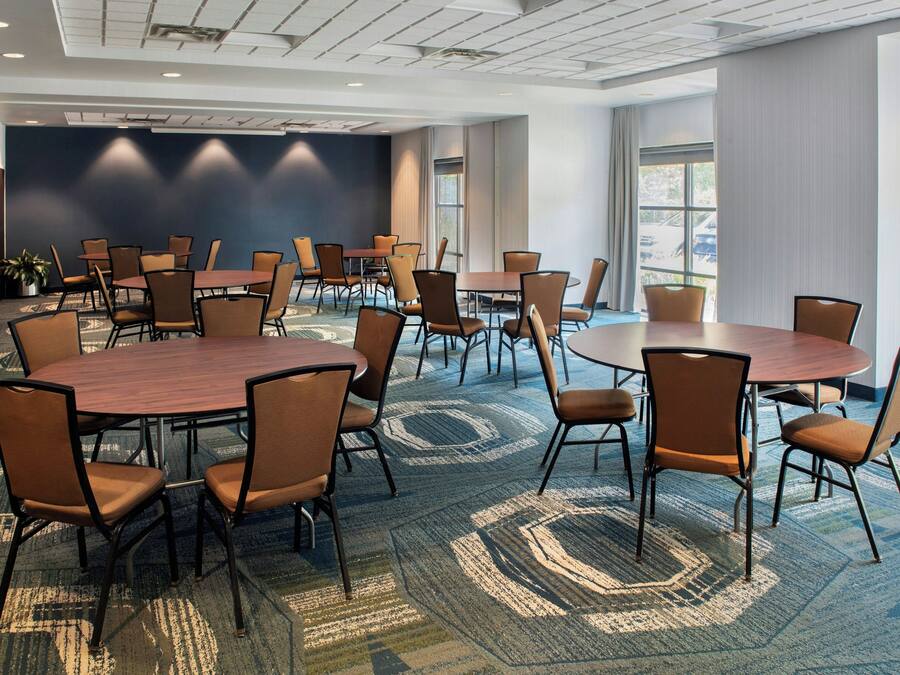 A conference room with multiple tables and chairs, carpeted flooring, and a large window providing natural light.