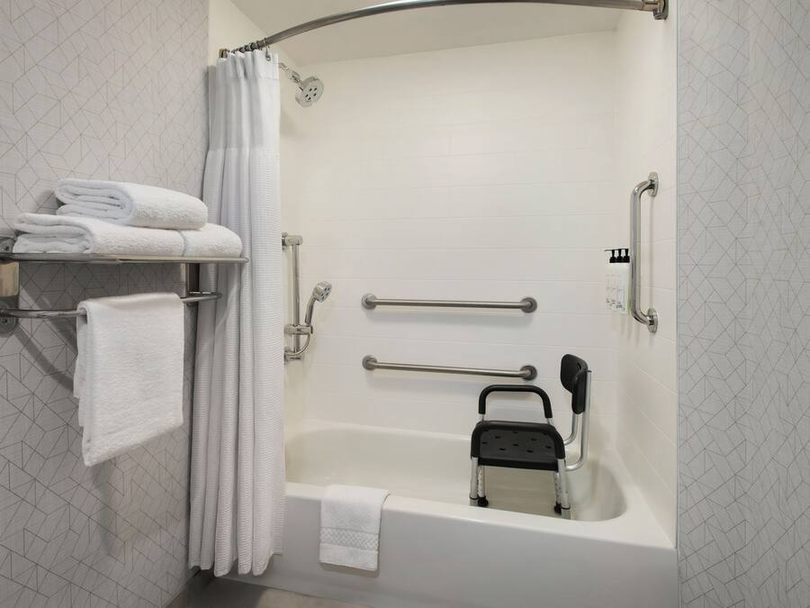 Un baño moderno con bañera, barras de apoyo, asiento de ducha y toallas ordenadas en un estante.