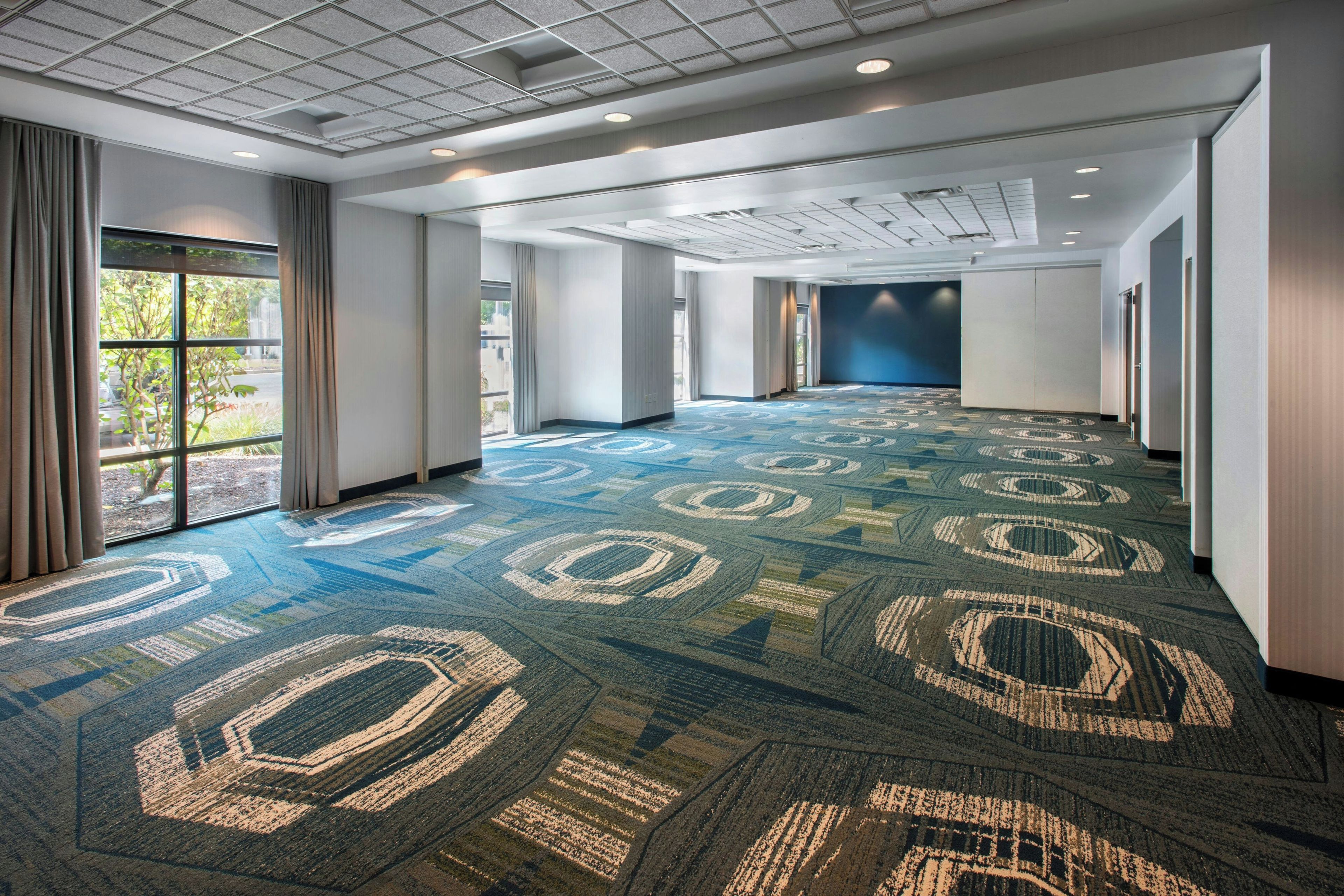 A spacious hotel corridor with patterned carpet, large windows, and a modern ceiling design.