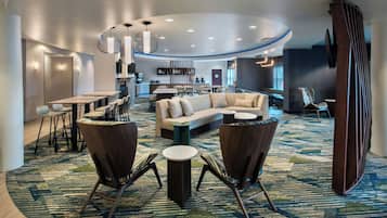 Un hall d’hôtel moderne avec un plafond incurvé, un espace salon avec des canapés et des fauteuils, une cuisine en arrière-plan et un tapis à motifs.