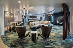 A modern hotel lobby with a curved ceiling, a seating area with sofas and chairs, a kitchen in the background, and a patterned carpet. - Springhill Suites by Marriott Pittsburgh Mills (Tarentum)