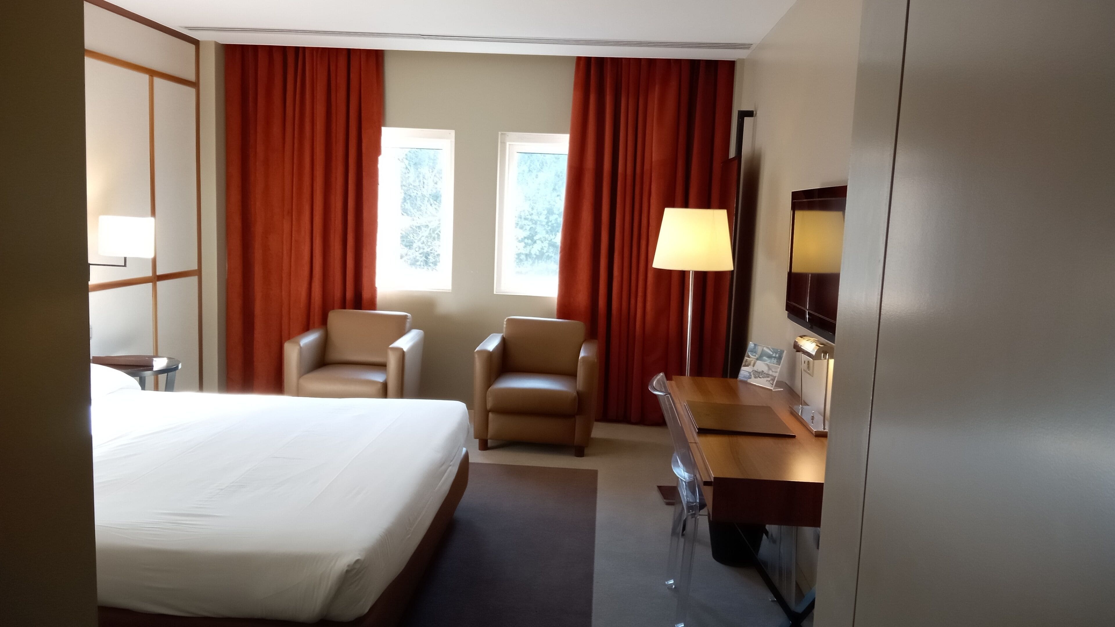 A hotel room with a bed, two chairs, a desk with a lamp, a TV, and a window with curtains.