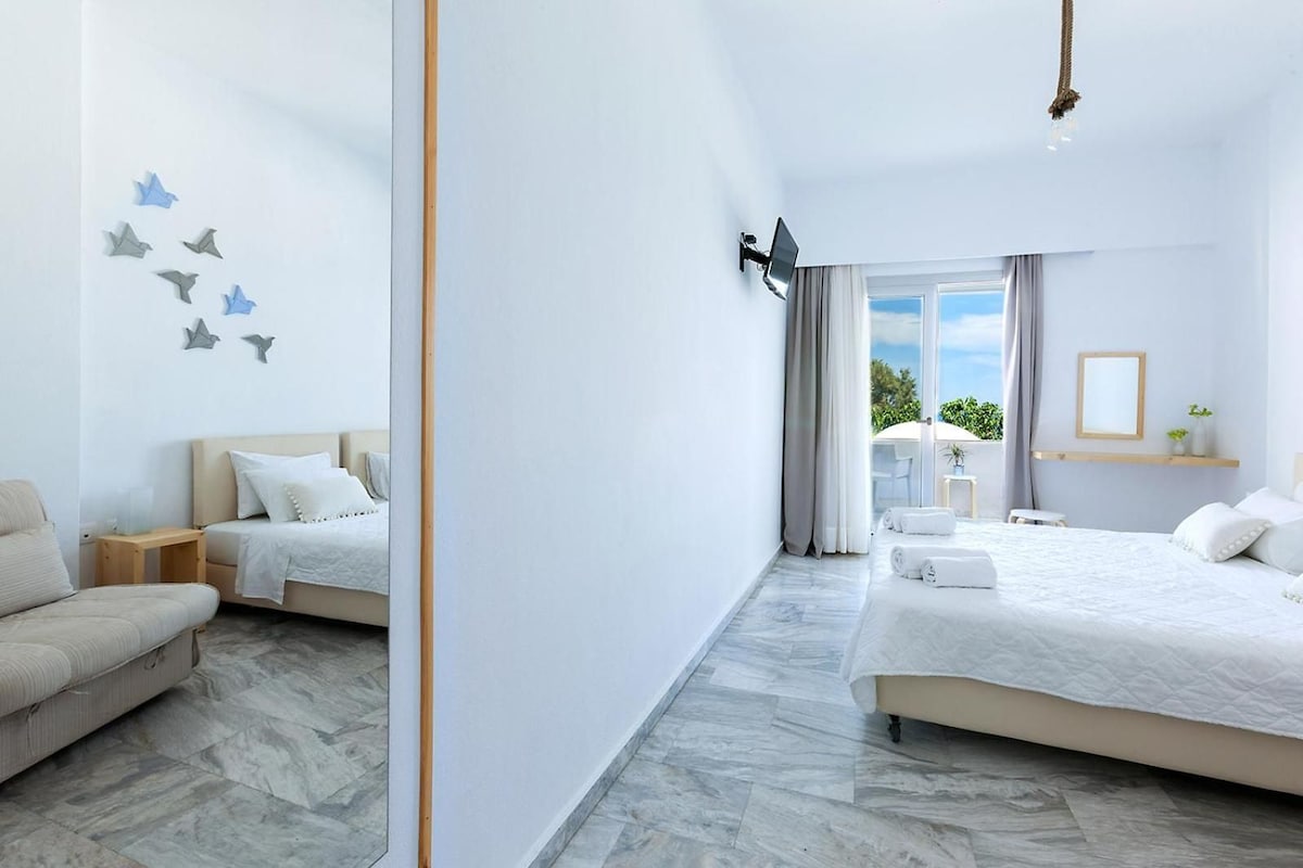 Quarto de hotel com uma cama grande, uma cama menor, um sofá, uma mesa de cabeceira, um espelho e uma janela com cortinas.