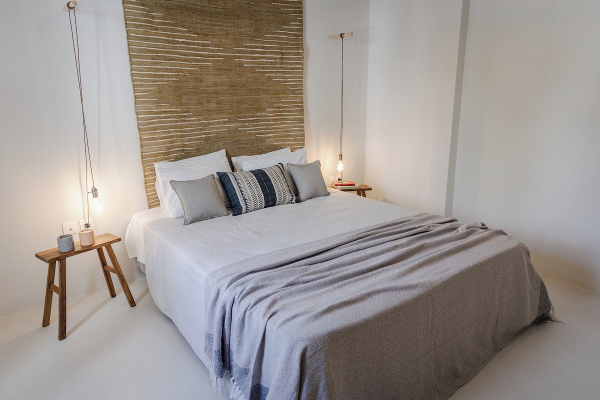 Una cama con cabecera de madera, dos mesitas de noche con lámparas, y una mesita de noche con un libro.