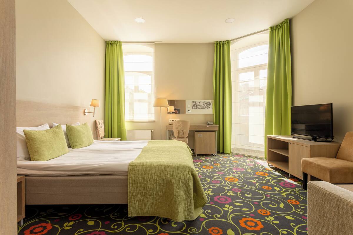 A hotel room with a bed, a desk, a chair, a television, and a carpet with a floral pattern.