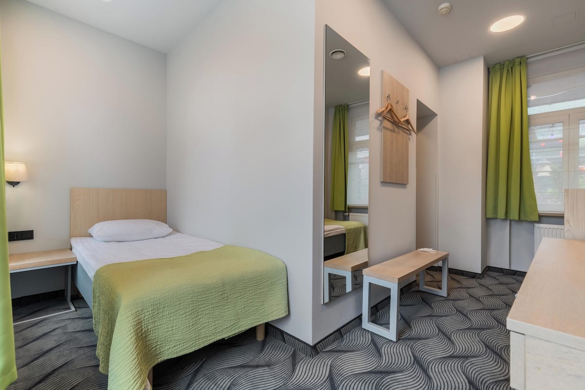 A modern hotel room with a single bed, a small bench, a mirror, and a window with green curtains.