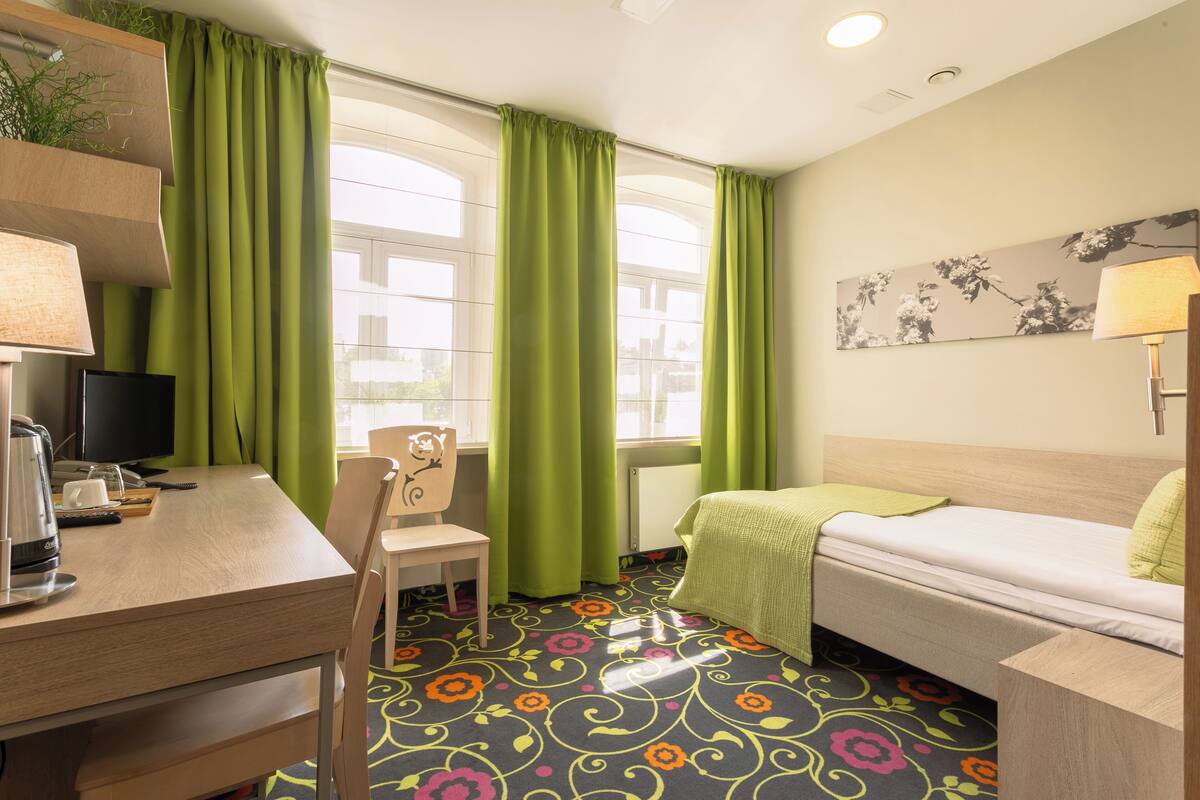 A hotel room with a bed, a desk, a chair, a TV, and a window with green curtains.