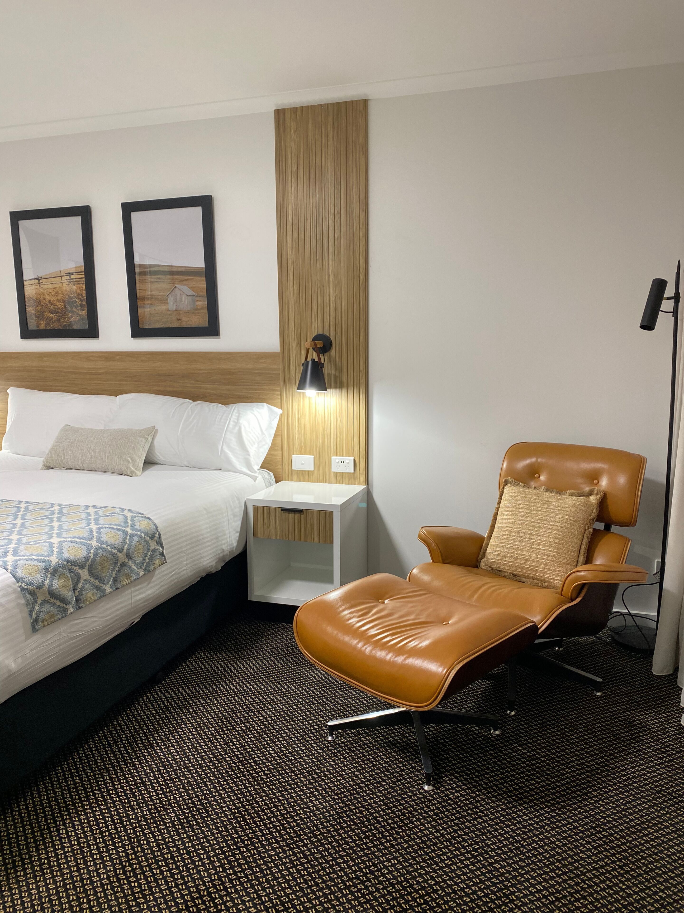 Habitación de hotel con cama, mesita de noche, lámpara, sillón de cuero y dos cuadros en la pared.