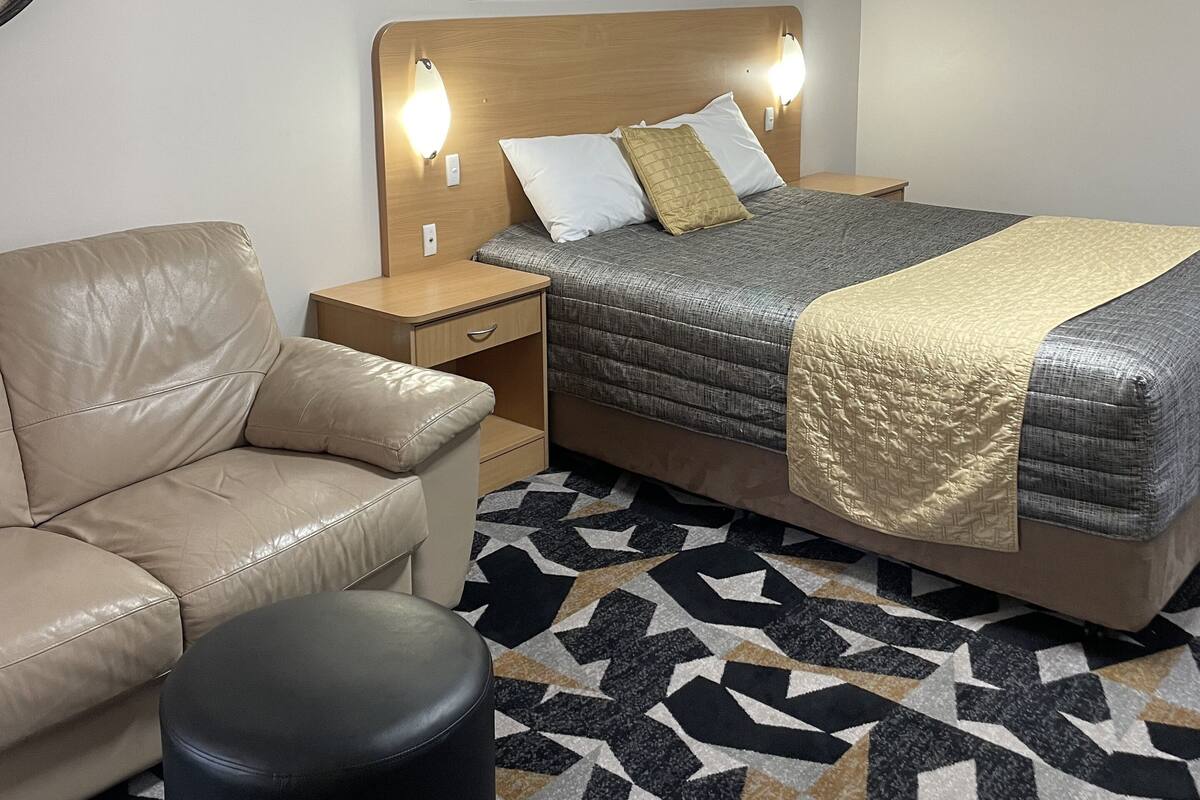 Une chambre d’hôtel comprenant un lit, une table de chevet, un canapé en cuir, un pouf noir, un tapis à motifs et une horloge murale.