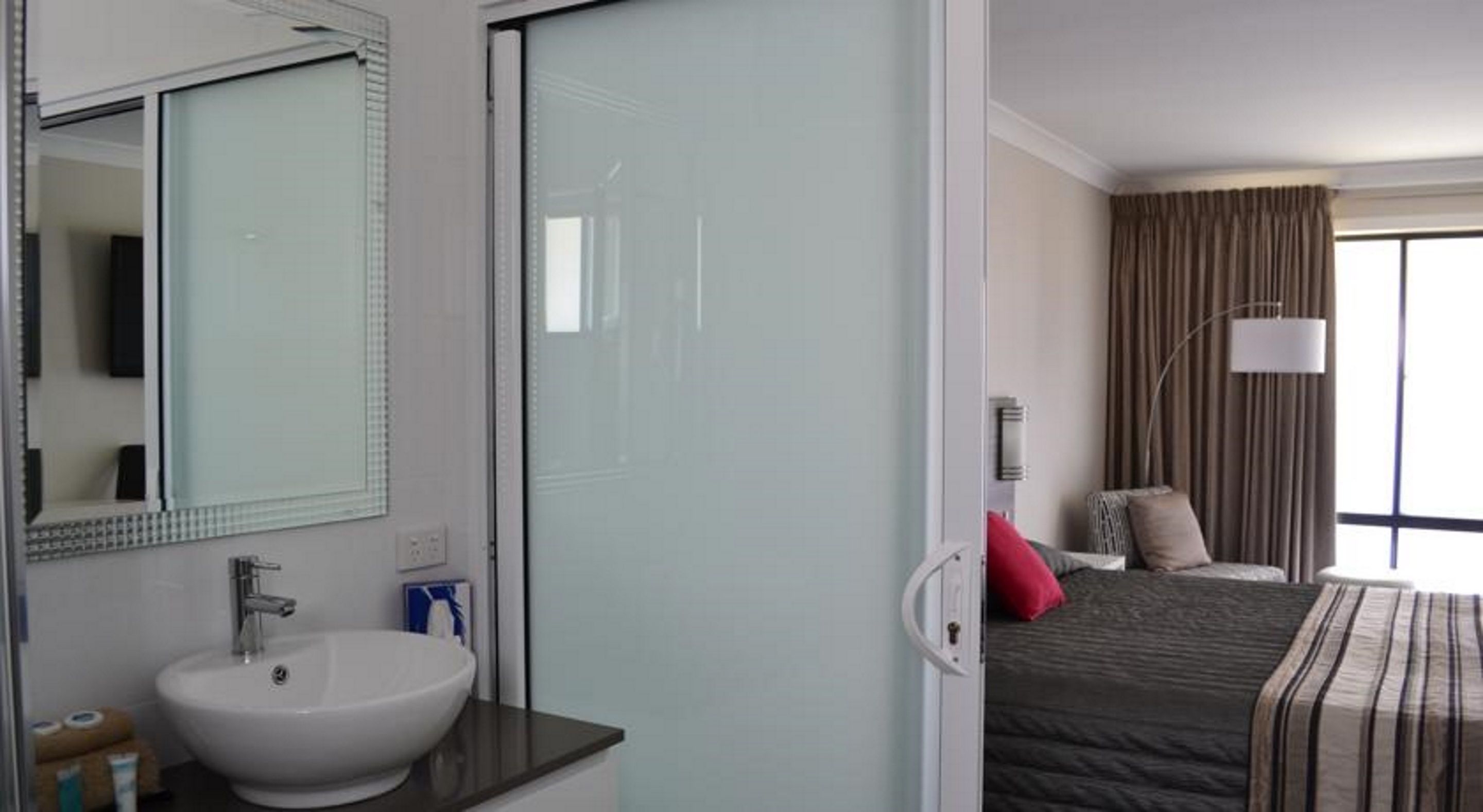 A hotel room with a large bed, a bedside lamp, a window with curtains, and a bathroom with a sink and mirror.