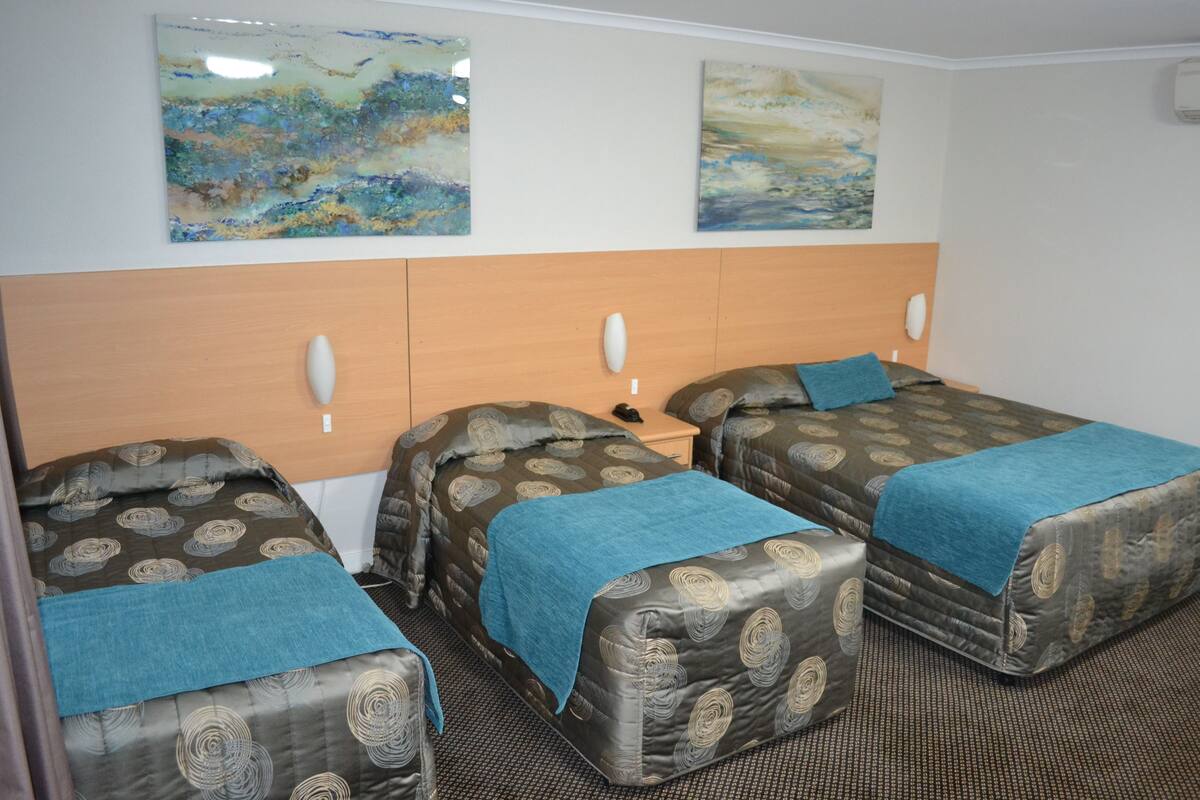 Une chambre d’hôtel avec trois lits simples, chacun recouvert d’une couverture bleue et d’une couette à motifs. Deux tableaux abstraits sont accrochés au mur, au-dessus des lits.