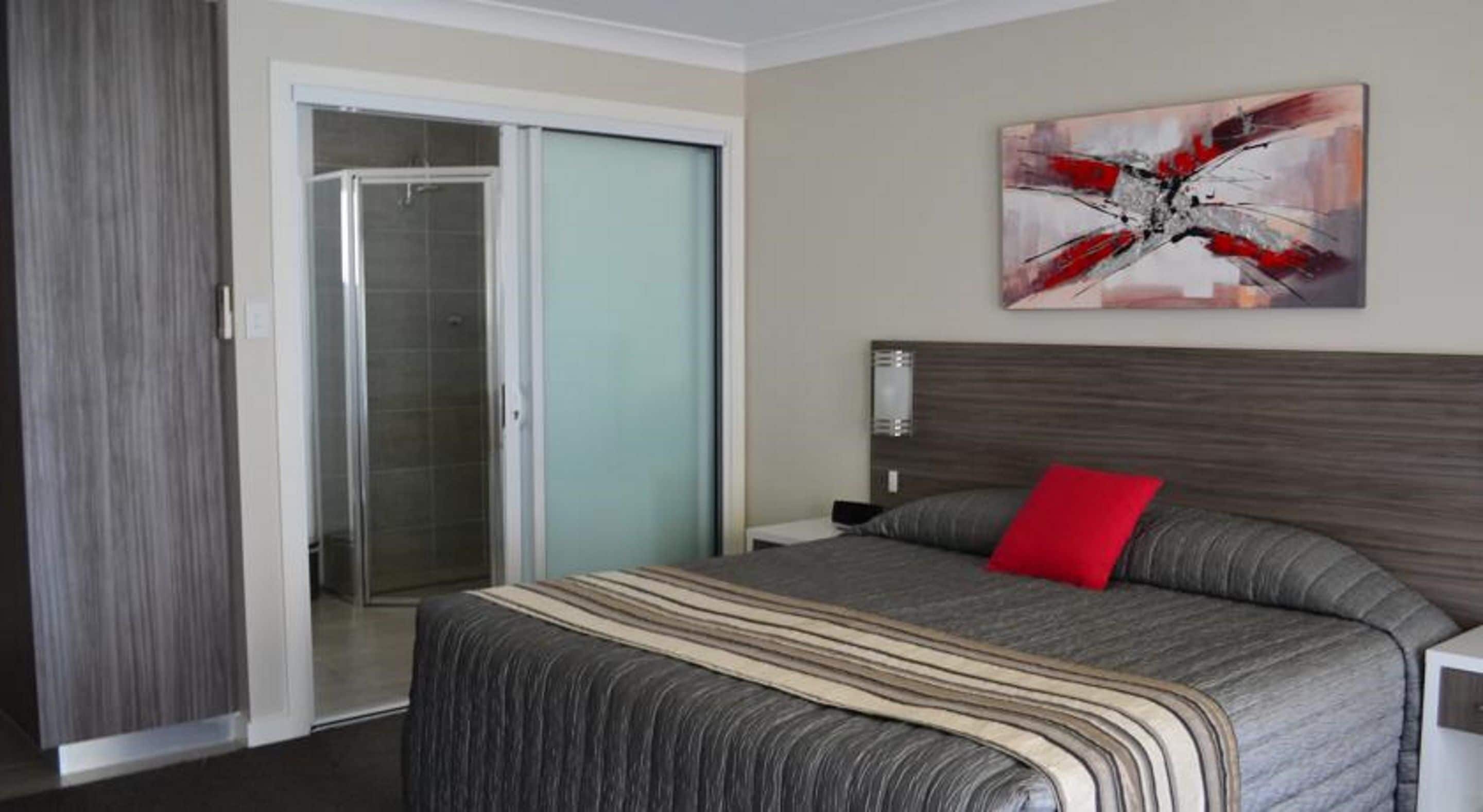 A bedroom with a bed, a red pillow, a shower, and a painting on the wall.