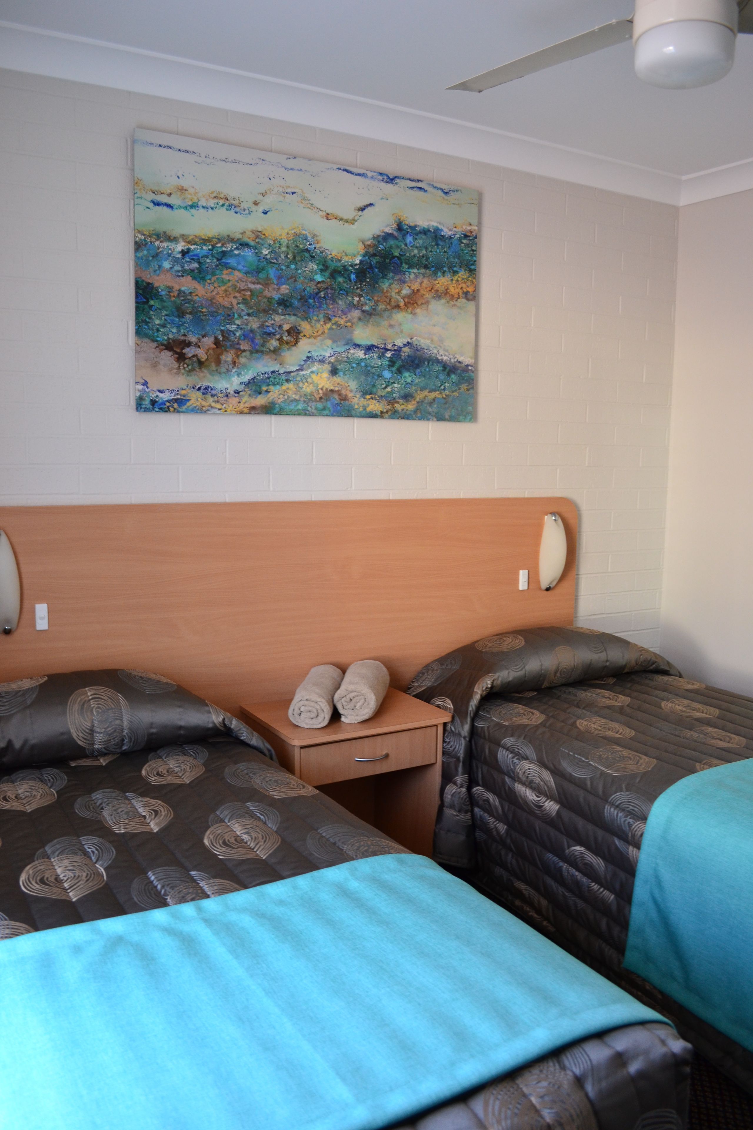 Habitación de hotel con dos camas, cabecera de madera, mesita de noche con toallas y un cuadro en la pared.