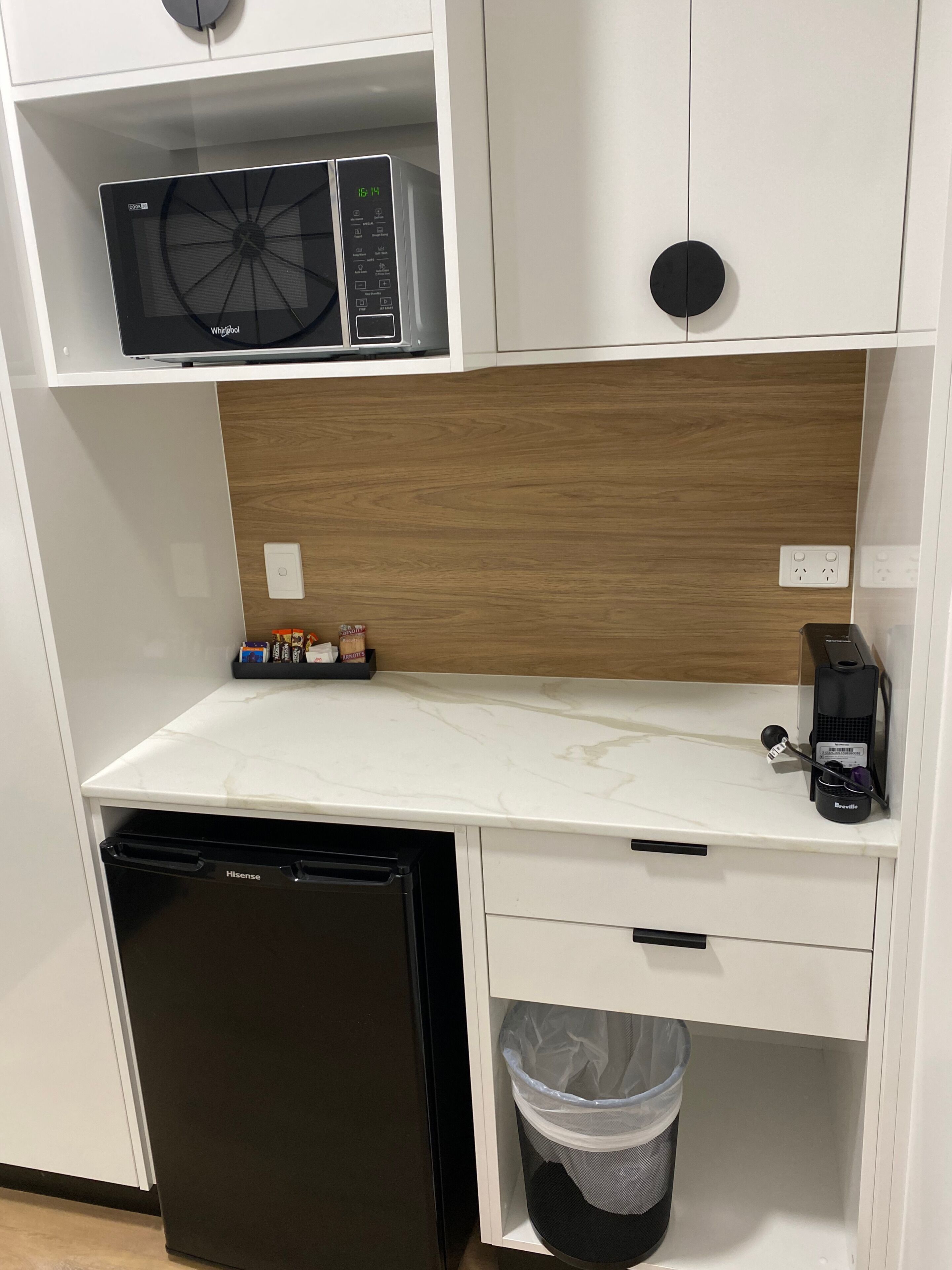 Een kleine kitchenette met een magnetron, koffiezetapparaat en een prullenbak.