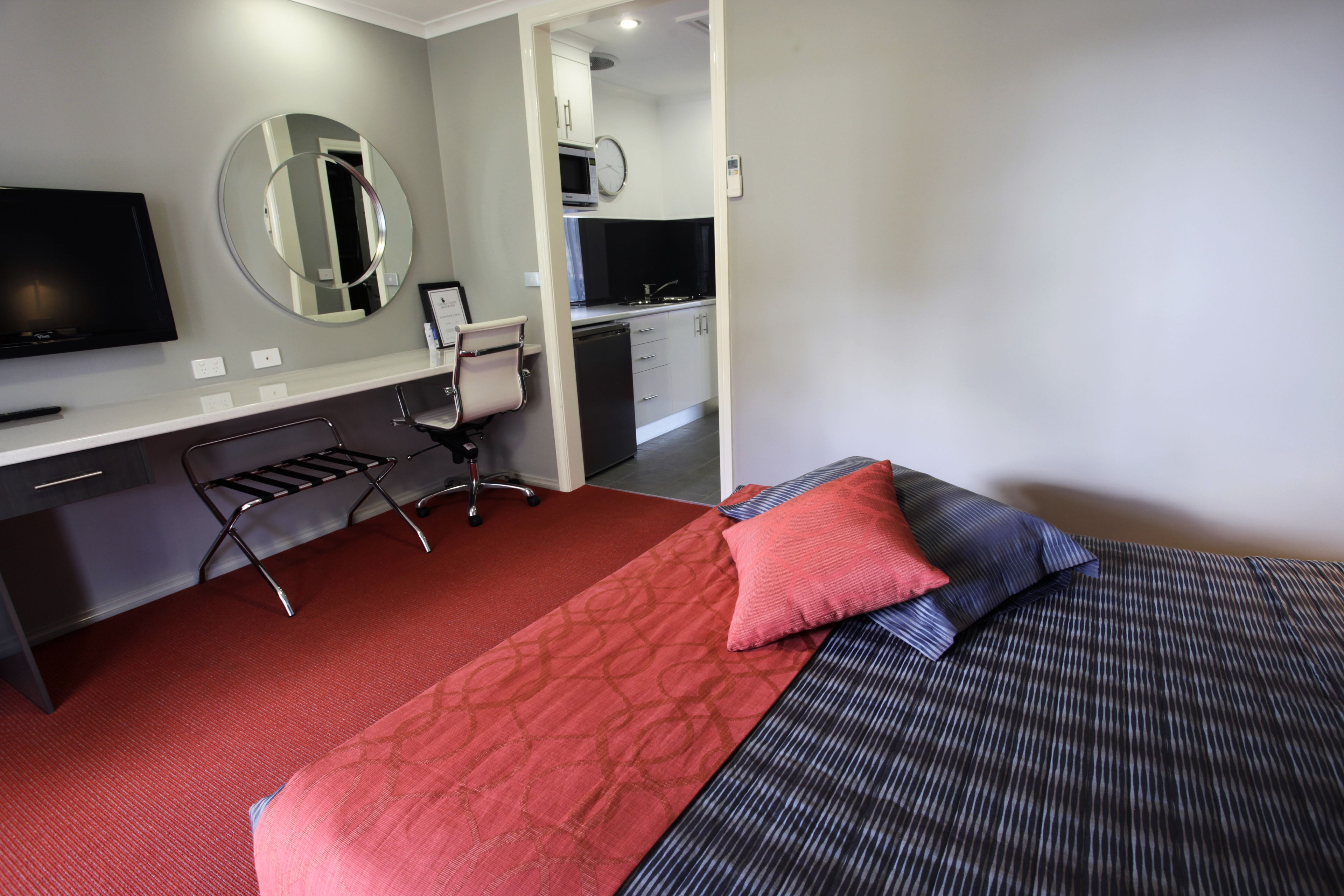 A hotel room with a bed, a desk with a chair, a TV, a mirror, and a kitchenette.