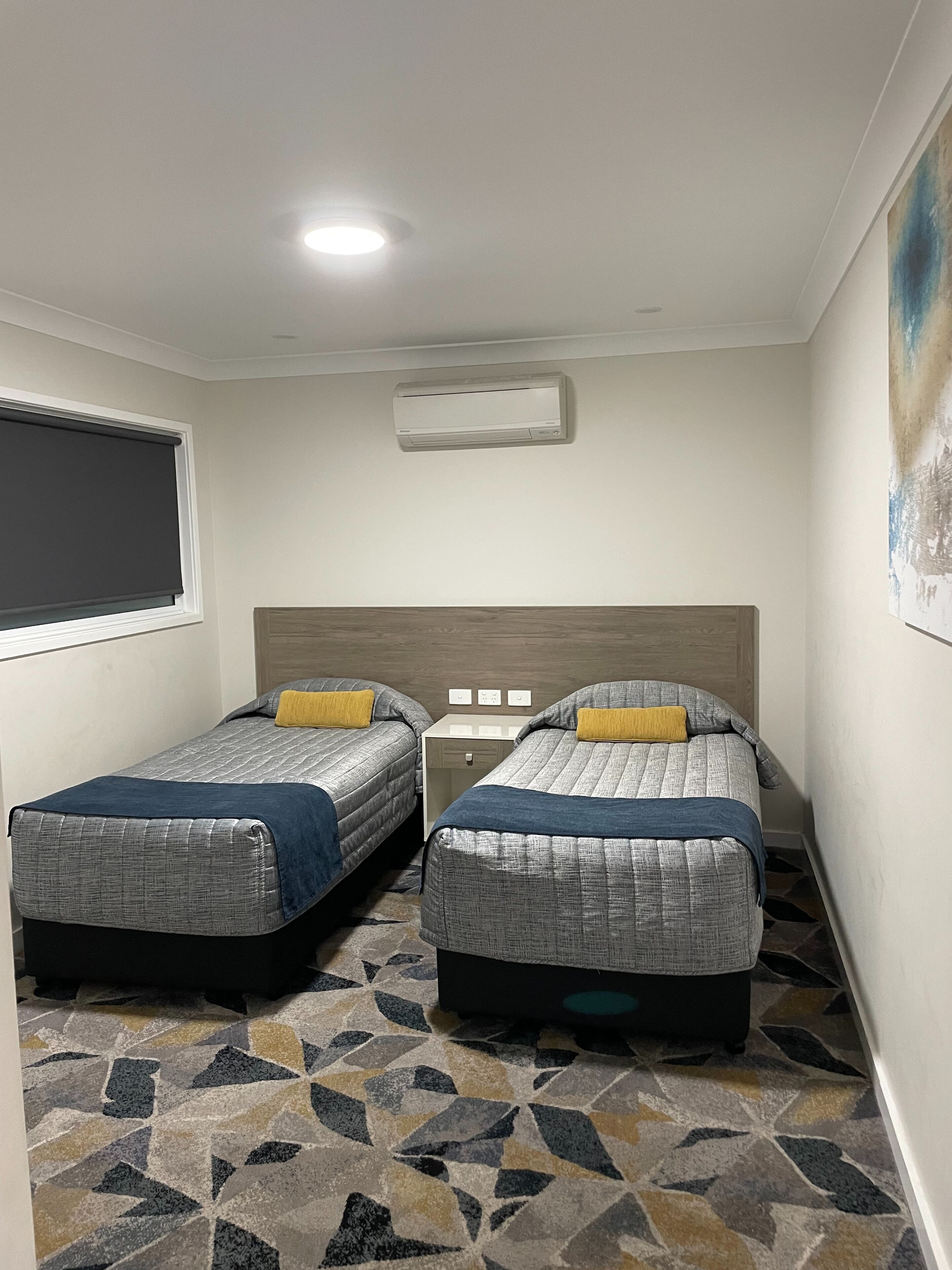 A small, compact room with two single beds, a window with a blind, a ceiling light, and a wall-mounted air conditioner.