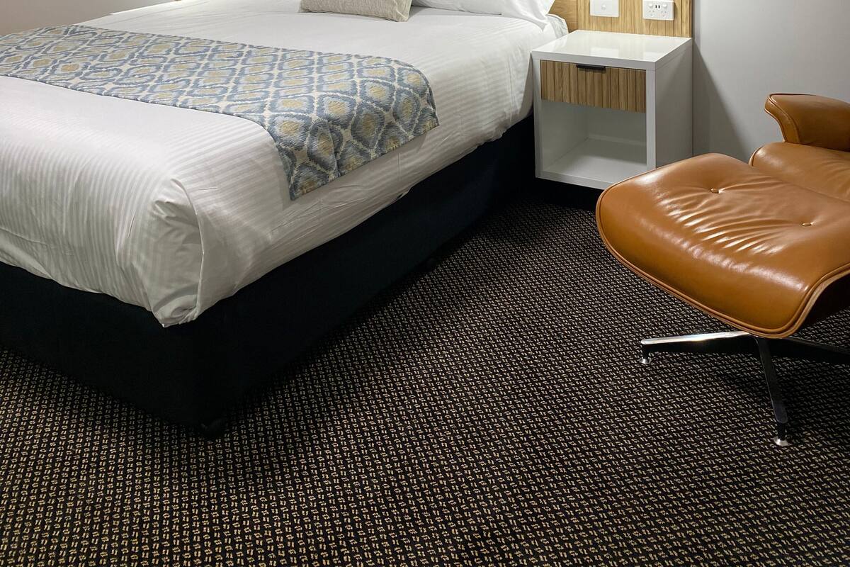 Une chambre d’hôtel avec un grand lit, deux tables de chevet avec des lampes, un fauteuil en cuir et une horloge murale.
