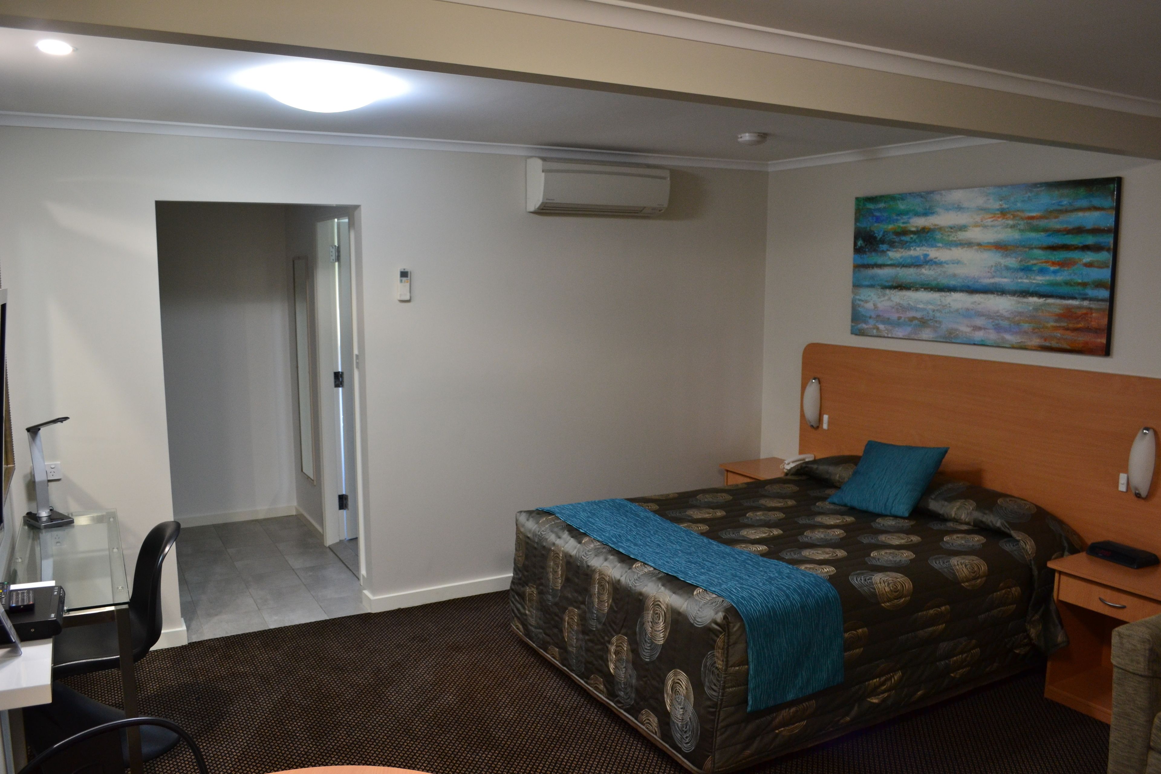 A hotel room with a bed, a desk, a chair, an air conditioning unit, and a painting on the wall.