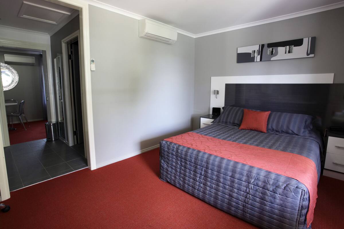 Une chambre d’hôtel avec un grand lit, une table de chevet, une commode, un climatiseur fixé au mur et une porte donnant sur une autre pièce.