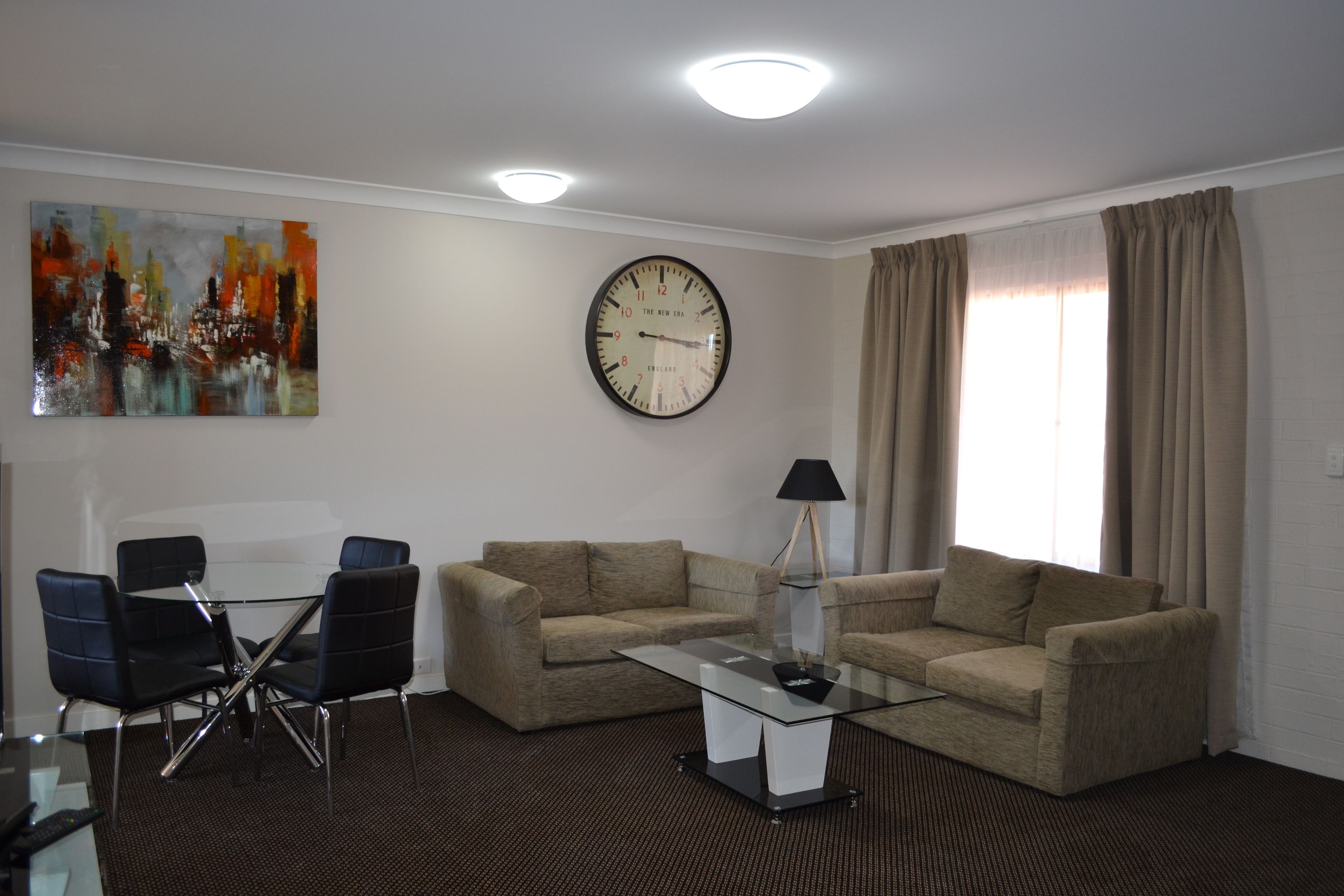 Uma sala de estar moderna com dois sofás, uma mesa de centro de vidro e uma área de jantar com cadeiras. Um relógio de parede grande e um quadro são visíveis.