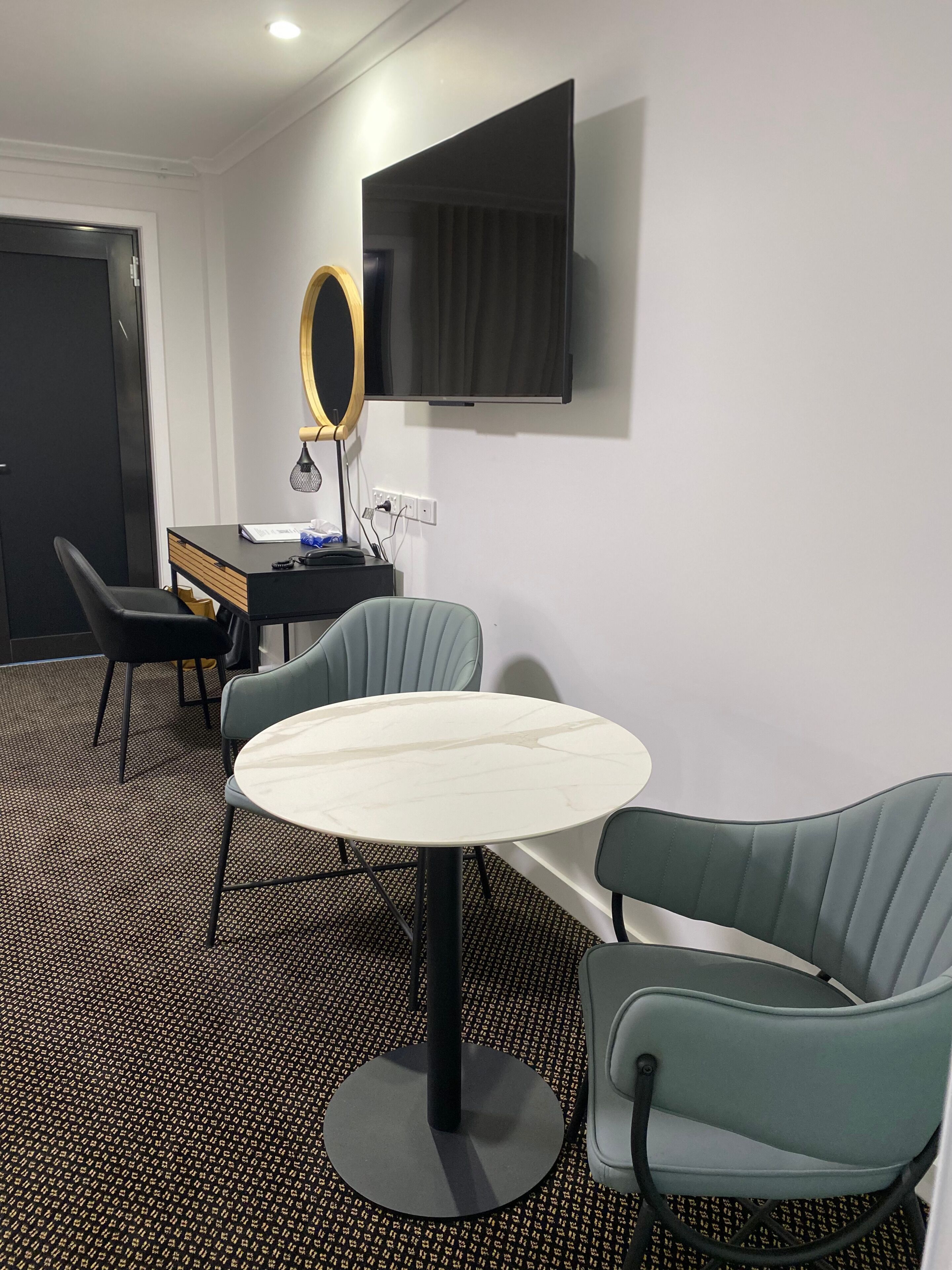 Quarto de hotel com uma mesa redonda, duas cadeiras, uma escrivaninha, um espelho e uma TV fixada na parede.