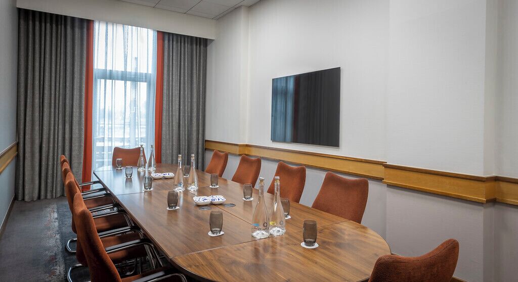 A conference room with a long wooden table, brown leather chairs, and a large window with grey curtains.