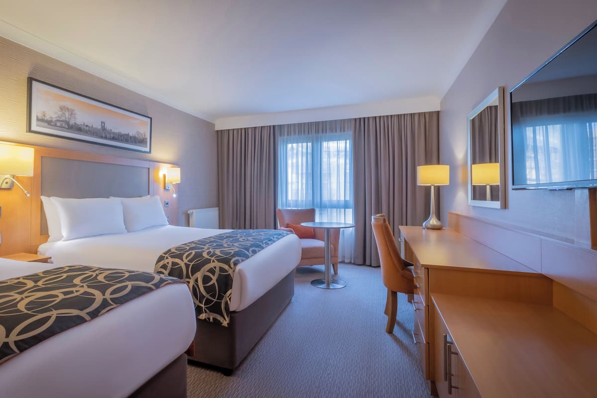 A hotel room with two beds, a desk, a chair, a small table, and a television.