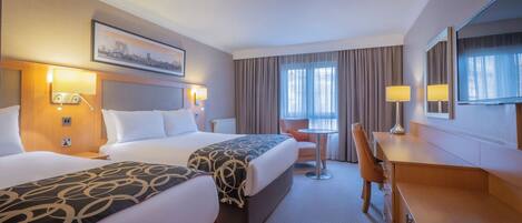 A hotel room with two beds, a desk, a chair, a small table, and a television.